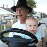 A candid photo of Gary driving a golf cart with his granddaughter Murphie in his lap. Murphie is a blonde toddler with brown eyes, and is wearing a blue and white striped shirt.