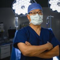 Dr. Joseph Kim, an East Asian man, stands with his arms crossed and poses for the camera while standing in an operating room. He is wearing blue UK scrubs, glasses, a face mask, and a surgical cap.
