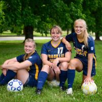 The three girls together crouching down on the field, each with a soccer ball, smiling.