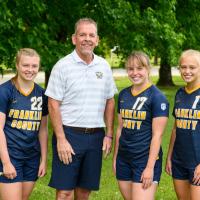 The three girls pose smiling with Coach Jon Sutphin, a tall middle aged man with a beard.