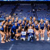 Izzy and Dalton hold signs with 10's on them and kneel in front of a group of UK gymnasts.