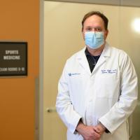 A portrait of Dr. Scott Mair, a middle-aged white man with brown hair and a white lab coat. He is standing next to a door labeled Sports Medicine Exam Rooms and is wearing a blue surgical mask.
