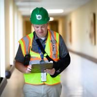 Leonard walks down a hall with a clipboard while wearing a safety vest and construction helmet.