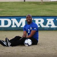 Remilson sits on the soccer field beside a soccer ball. He is wearing his blue number seven Danville soccer jersey with black pants and black and white sneakers.