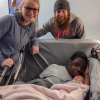 Remilson lays in bed next to his newborn niece. His sister Michaela, a young woman with long blonde hair and her husband, a young man with a red long beard, stand beside the bed smiling.
