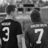 A black and white photo of Zach and Remilson, smiling and talking to each other. Their backs are to the camera and we can see the back of their soccer jerseys, with number three (Zach) and number seven (Remilson).
