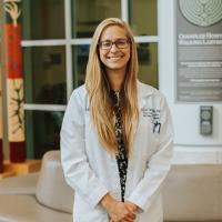 A photo of Dr. Maya Ignaszewski holding her hands in front of her as she smiles for the camera. She is a white woman with long blonde straight hair. She is wearing a white lab coat over a black shirt with a white pattern on it.