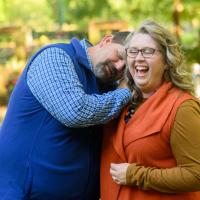 A candid photo of Ronald leaning his head on his wife, Jessica’s, shoulder as she laughs. She is a white woman with long curly blonde hair. She is wearing an orange vest over a long-sleeve brown sweater, and a pair of dark-rimmed glasses.