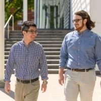 Ronnie and his roommate Zack, a young white man with long brunette hair, smile as they walk together outside. Zack is wearing a silver watch, round dark-framed glasses, and a white button-up shirt that is tucked into his khakis.