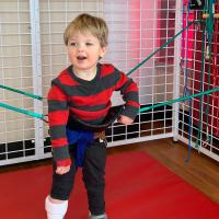 Rory at physical therapy in his gait trainer. He is wearing a dark grey and red striped shirt with dark grey pants.