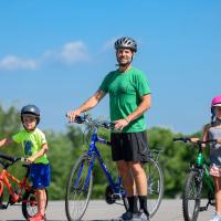 Ryan, wearing a helmet, stands next to a blue bicycle. He is flanked by his young son and daughter, who are both standing by their bicycles and wearing helmets.