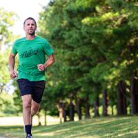 Ryan runs along a road next to a clump of trees. He is wearing a green shirt and black shorts.