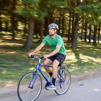 Ryan rides his bike along a road next to a clump of trees. The background is blurred slightly. He is wearing a green shirt and a helmet.