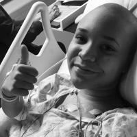 Black and white photo of Savannah laying in a hospital bed smiling with a thumbs up.