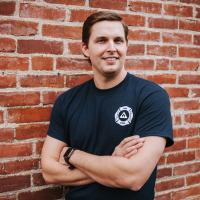 Trent stands in front of an exposed brick wall with a smile and his arms folded. He is wearing a navy blue EMS rescue tee shirt, and his brown hair is slicked back.