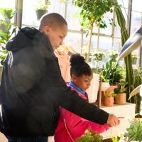 A photo of Whitley and Essence in a greenhouse. Both of their hands are extended and reaching out to touch the leaves of a plant.